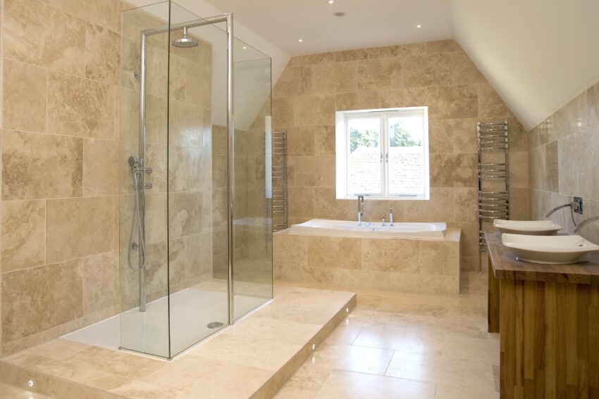Большая ванная комната со стеклянной душевой кабиной, полом из травертина и двойной раковиной на деревянной столешнице.