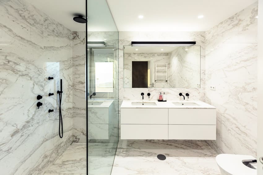 Ванная комната со стенами из каррарского мрамора, стеклянной перегородкой, насадкой для душа, парящим туалетным столиком, зеркалом и раковиной.