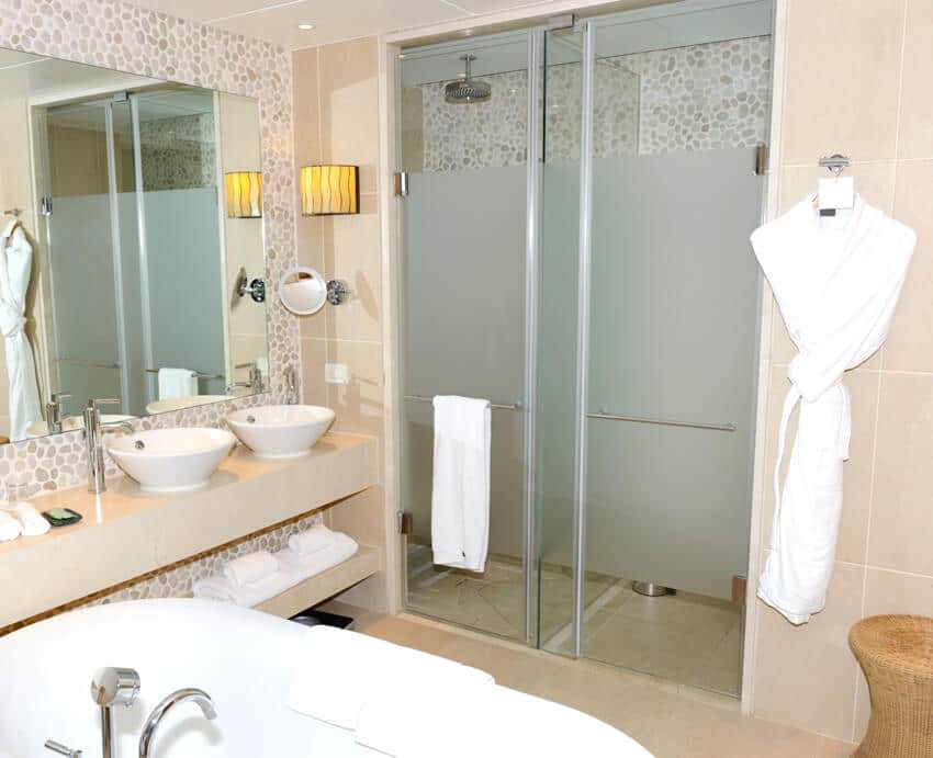 Интерьер ванной комнаты с ванной, двумя раковинами и душевыми дверями из частично матового стекла с халатом и полотенцем рядом с ним
