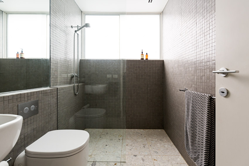 Ванная комната с душевой стеной из фарфоровой мозаики, насадкой для душа, держателем для полотенец, окном, унитазом и зеркалом.