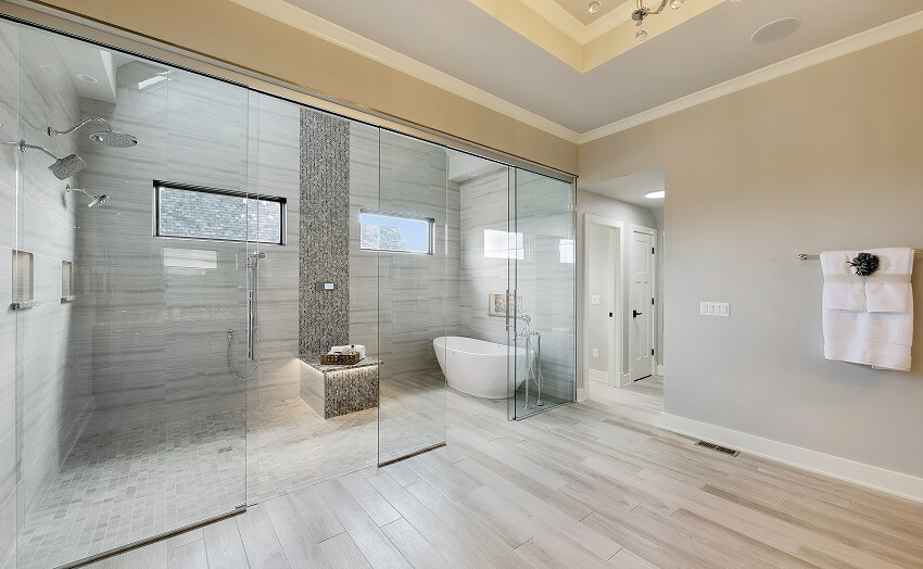 Современная ванная комната с отдельно стоящей ванной, двойным душем, стеной из серой плитки и скамейкой из мозаичной плитки, заключенной в стекло