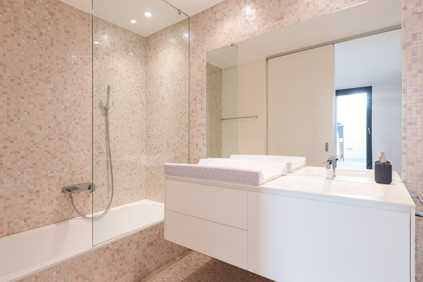 Ванная комната с туалетным столиком, зеркалом, столешницей, раковиной, душевой стеной, выложенной мозаичной плиткой, и стеклянной перегородкой.