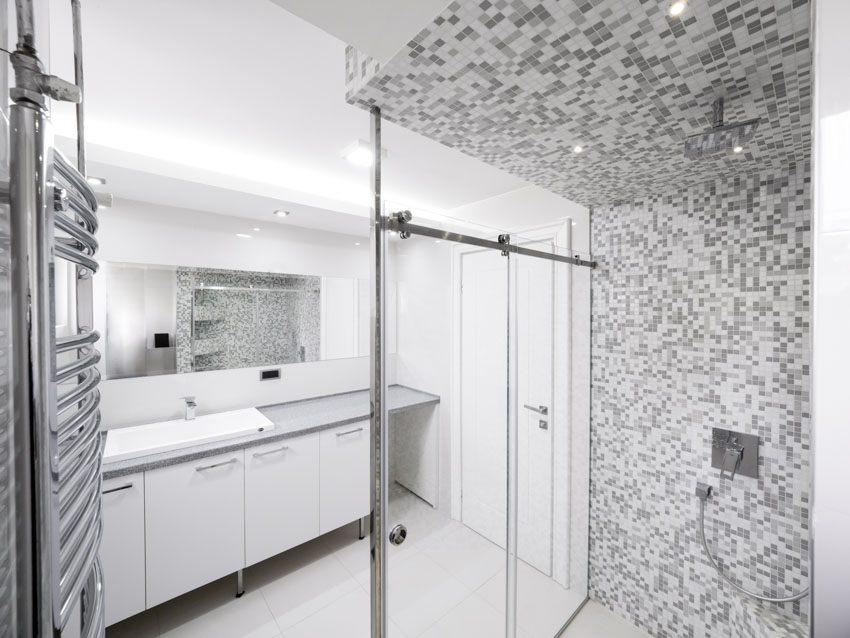 Ванная комната с мозаичной душевой стеной, стеклянной перегородкой, держателем для полотенец, косметическим зеркалом, столешницей, шкафчиками и раковиной.