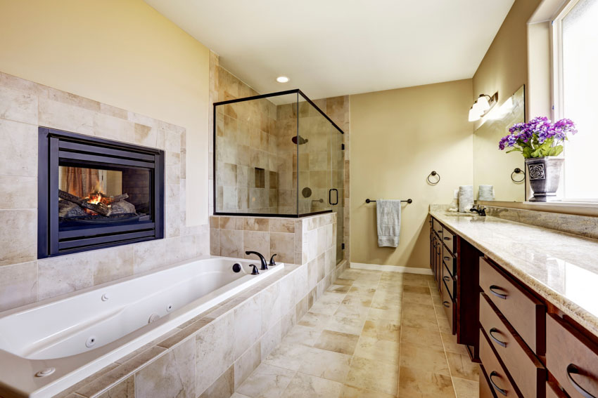 Ванная комната с камином, кафельным полом, ванной, столешницей, выдвижными ящиками, невысокой стеной и стеклянной душевой кабиной.