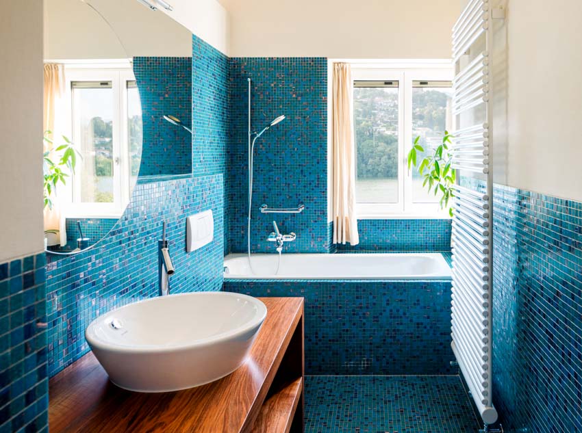 Ванная комната с душем из синей стеклянной плитки, деревянным туалетным столиком, раковиной, краном, зеркалом и окнами.