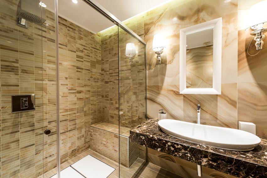 Ванная комната со столешницей, раковиной, зеркалом, настенными светильниками и душем из стеклянной плитки.