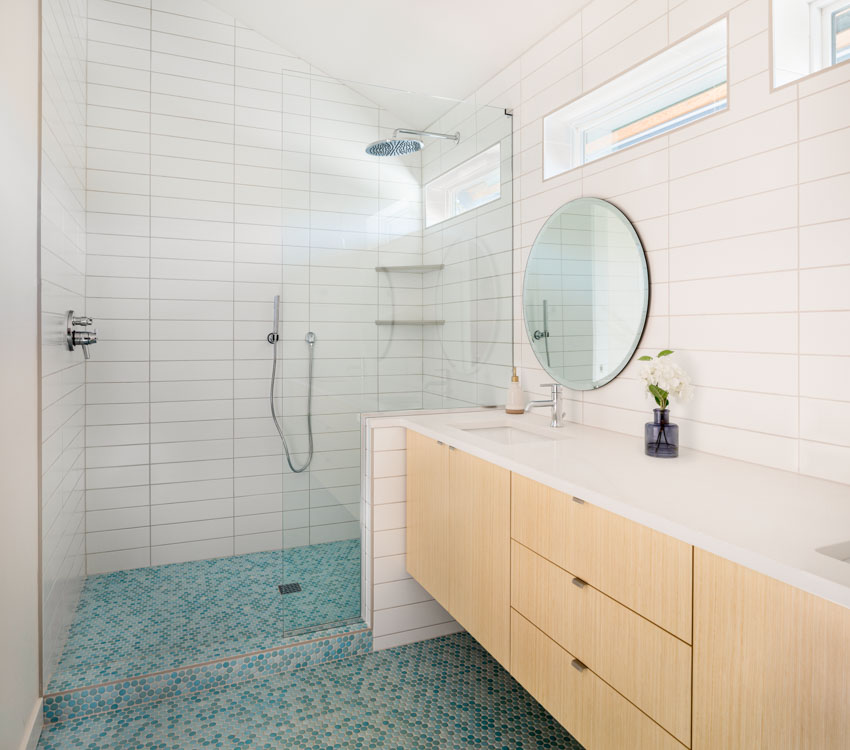Ванная комната со стеклянным плиточным полом, стеной из плитки метро, ​​стеклянной дверью, шкафами, столешницей, раковиной, зеркалом и окнами.