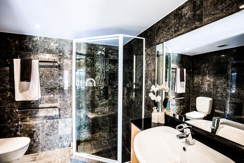 Ванная комната с гранитной душевой стеной, стеклянной дверью, зеркалом, раковиной, краном и держателем для полотенец.