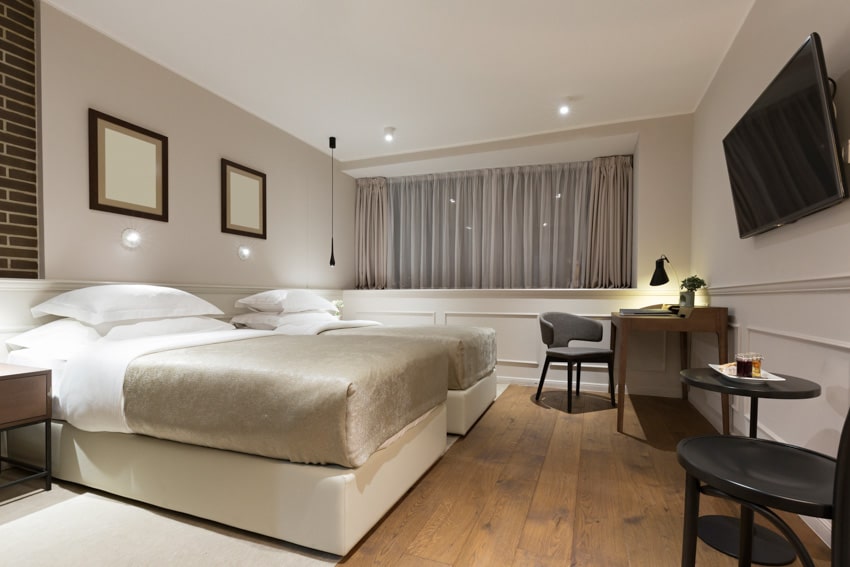 Спальня со встроенным настенным бра, постельным бельем, подушками, столом, стульями, деревянным полом, окном, шторами и телевизором.
