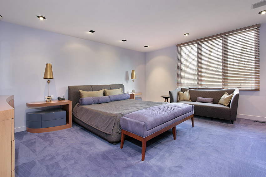 Простая спальня с тумбочкой, скамейкой, диваном, ковровым покрытием, окрашенной в лавандовый цвет стеной, лампой и окнами.