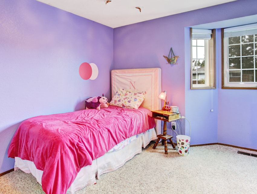Спальня с розовыми постельными принадлежностями, изголовьем, подушкой, эркером, тумбочкой и окрашенной в лавандовый цвет стеной