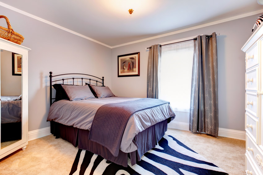 Простая спальня с подушками, постельным бельем, зеркалом, окном, шторами и окрашенной в лавандовый цвет стеной