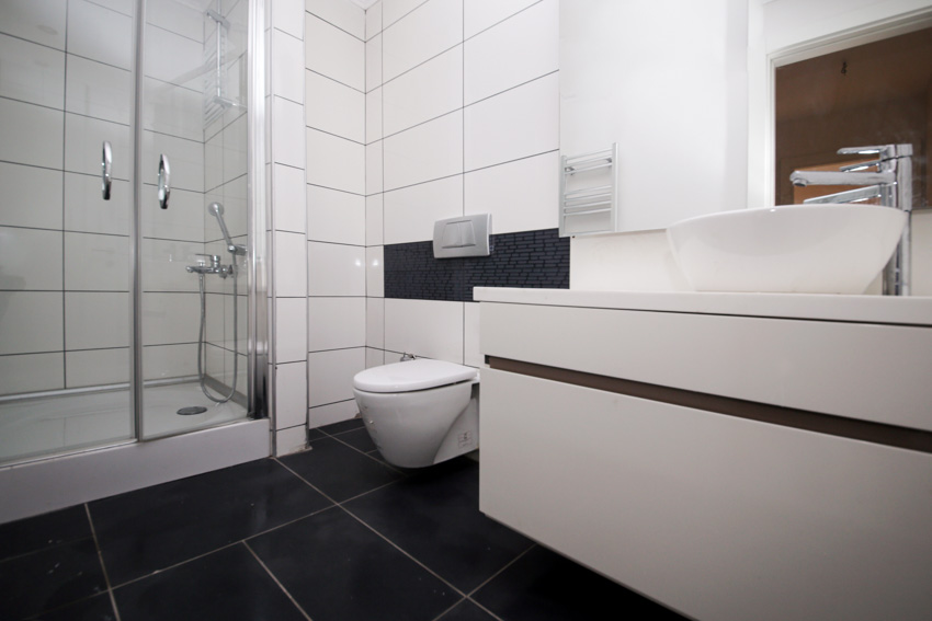Ванная комната с черной напольной плиткой разного размера, туалетным столиком, туалетом, душем, стеклянной дверью, краном и зеркалом.