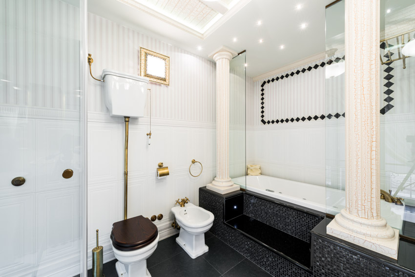 Ванная комната с черной напольной плиткой с золотой отделкой, туалетом, ванной, белыми стенами и колоннами.