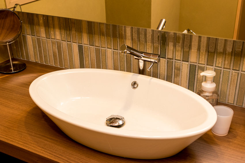 Ванная комната с зеркалом, раковиной, фартуком из мини-плитки, краном и деревянной столешницей.