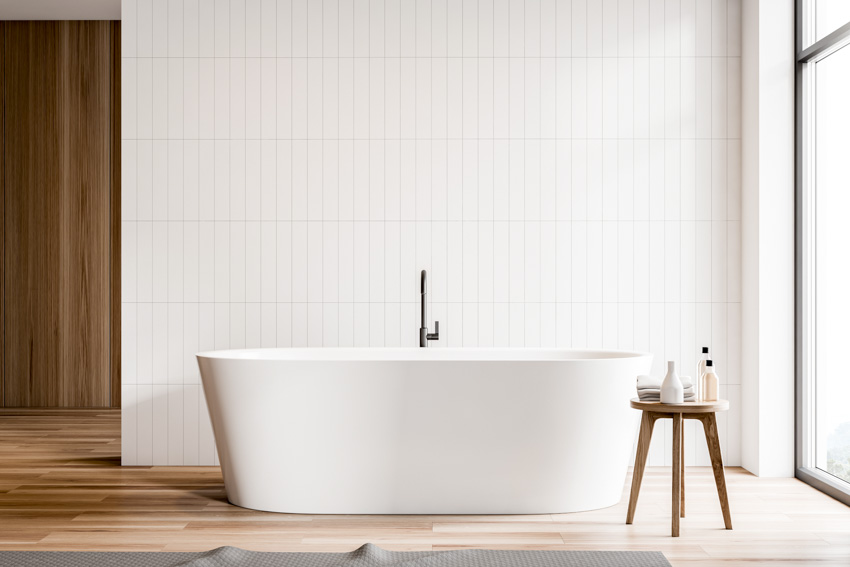Минималистская ванная комната с плиткой на стене, ванной, табуретом, окном и деревянным полом.