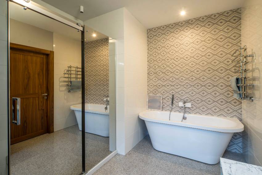Современный светлый интерьер ванной комнаты с плиточной акцентной стеной, деревянной дверью, белой ванной и зеркалом на стене
