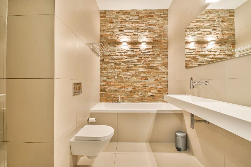 Белый кафельный интерьер ванной комнаты с каменной акцентной стеной, ванной и туалетом