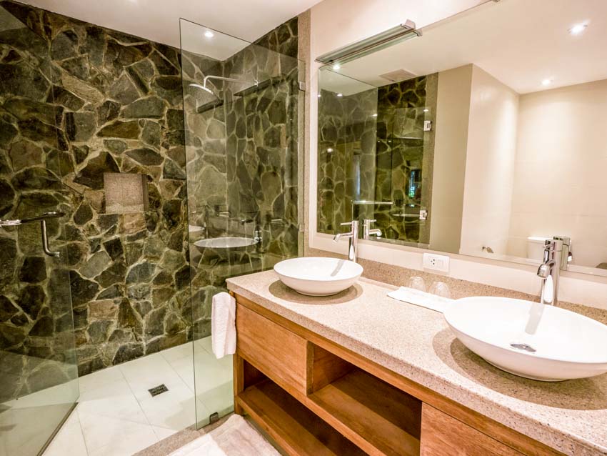 Ванная комната с каменной душевой стеной, стеклянной перегородкой, гранитной столешницей, раковиной, смесителем и зеркалом.