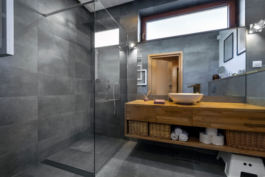 Ванная комната с крупноформатной плиткой, душем, стеклянным ограждением, плавающей деревянной столешницей, раковиной, зеркалом и окном.