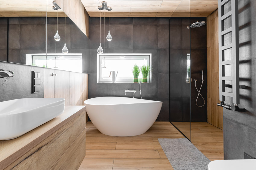 Ванная комната с черной крупноформатной плиткой, душем, ванной, стеклянной перегородкой, косметическим зеркалом, раковиной, деревянными шкафчиками и окнами.