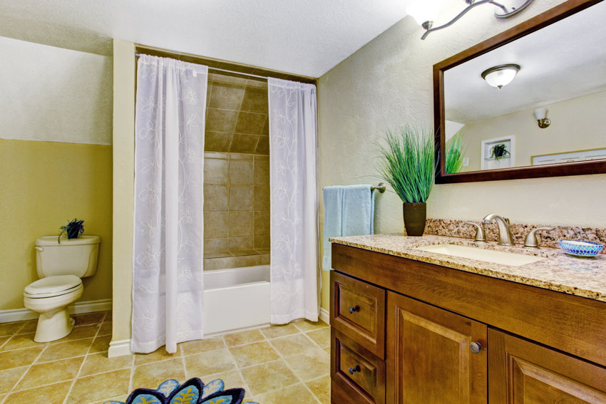 Ванная комната с прозрачной занавеской для душа, кафельным полом, туалетом, зеркалом, столешницей, шкафчиками и раковиной.