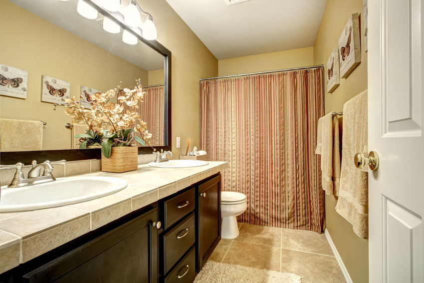 Ванная комната с занавеской для душа, туалетным столиком, ящиками, шкафом, столешницей, зеркалом и унитазом.