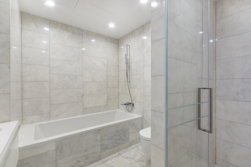 Ванная комната с ванной, насадкой для душа, стеной из каррарского мрамора, полом и стеклянной дверью.