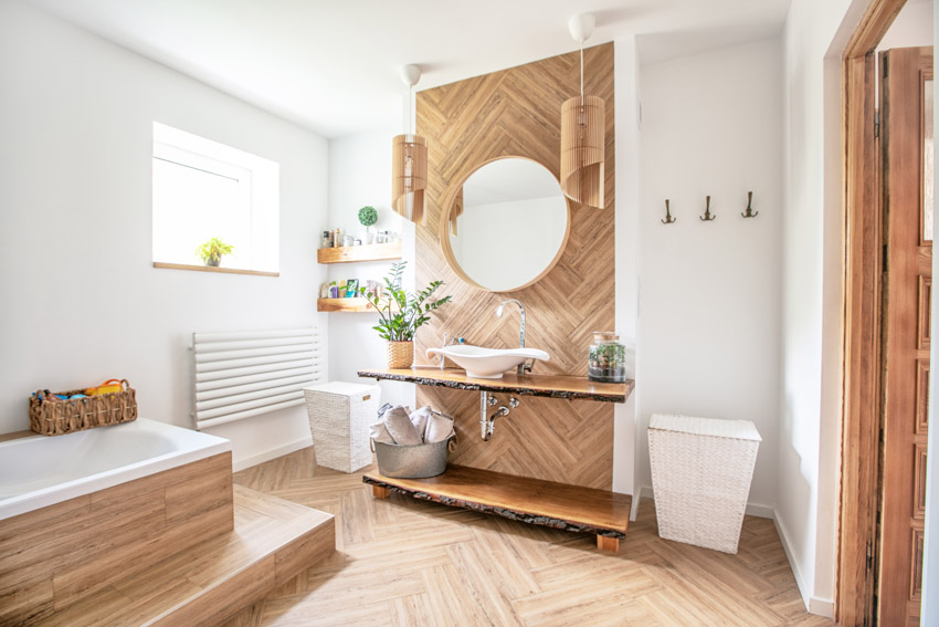 Красивая ванная комната с косметическим зеркалом, ванной, плавающей столешницей, раковиной, окном и деревянным полом.