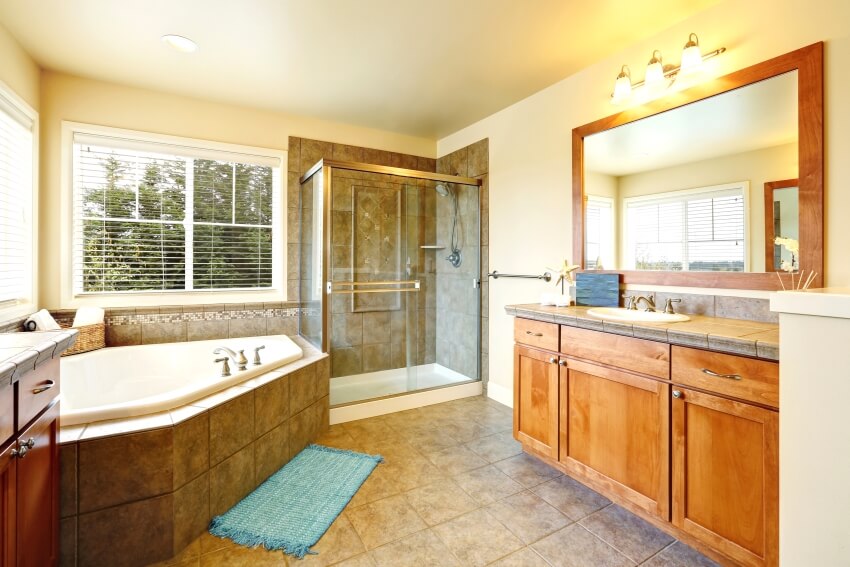 Ванная комната с двумя тумбочками, стеклянной душевой кабиной и угловой ванной, облицованной серо-коричневым мрамором.