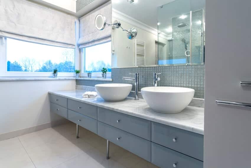 Современный интерьер ванной комнаты с двумя раковинами, фартуком из стеклянной плитки, мраморными столешницами и окнами