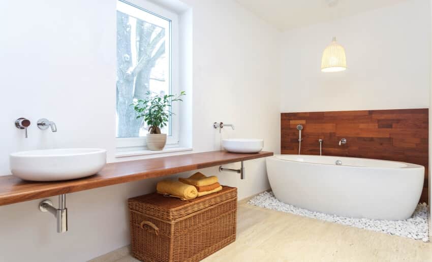 Белая ванная комната с отдельно стоящей ванной, местом для хранения корзин и двумя раковинами на парящей столешнице из деревянных плит.