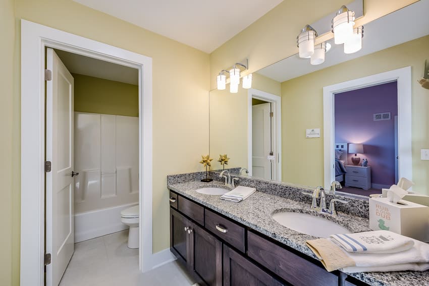 Ванная комната с бледно-желтыми стенами, гранитной столешницей, акцентным освещением, зеркалом и двумя раковинами.