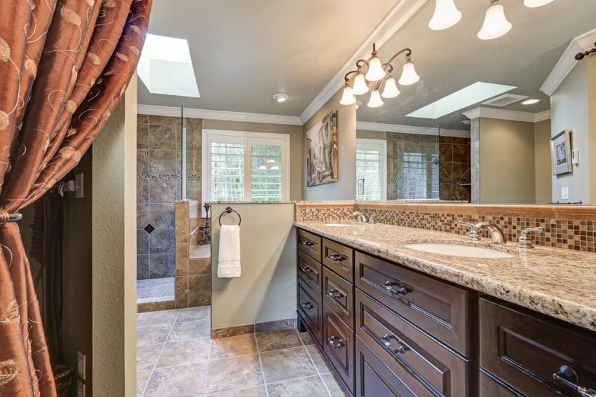 Роскошная ванная комната с гранитной столешницей, деревянными ящиками, акцентным освещением, зеркалом и потолочным окном.