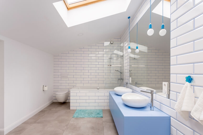 Мансардная ванная комната со стенами из плитки метро, ​​световым окном и умывальниками на столешницах, окрашенных в синий цвет.