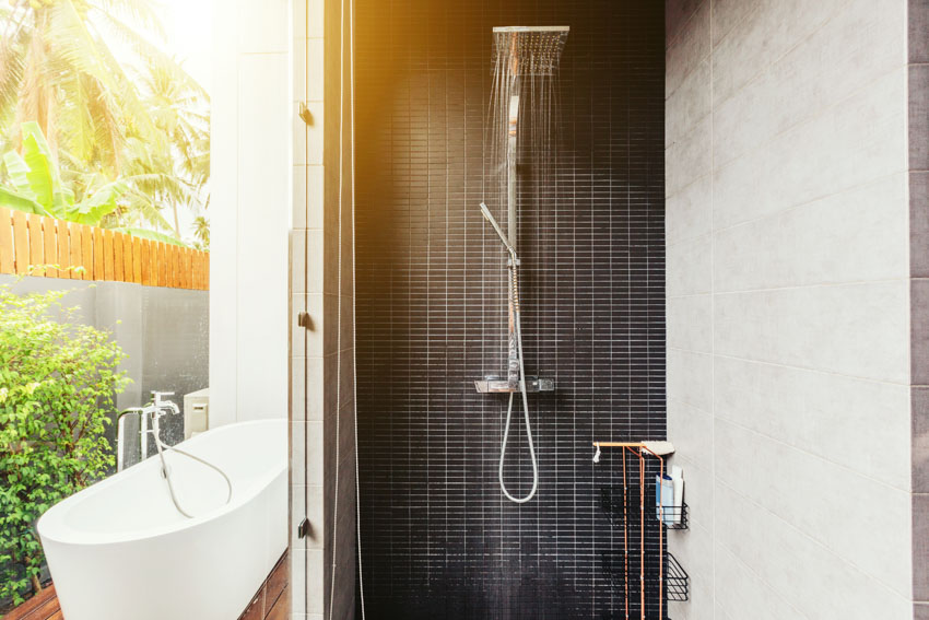 Ванна на открытом воздухе с ванной, современный душ, стена из черной плитки и растения