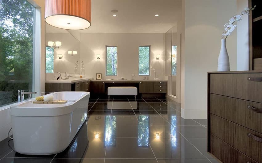 Современная главная ванная комната с отдельно стоящей черной ванной, гранитными полами и белыми туалетными столиками.