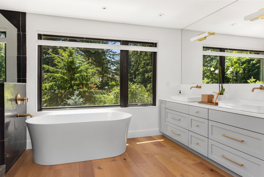 Ванная комната с акриловой ванной, ящиками, зеркалом, окнами и деревянным полом.