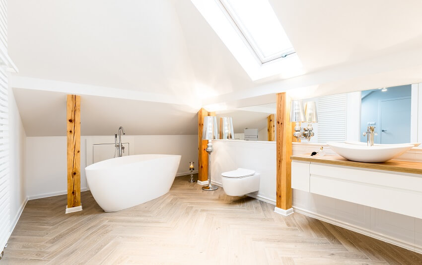 Белая ванна и туалет в просторной мансардной ванной комнате с окном в крыше, балками и зеркалом