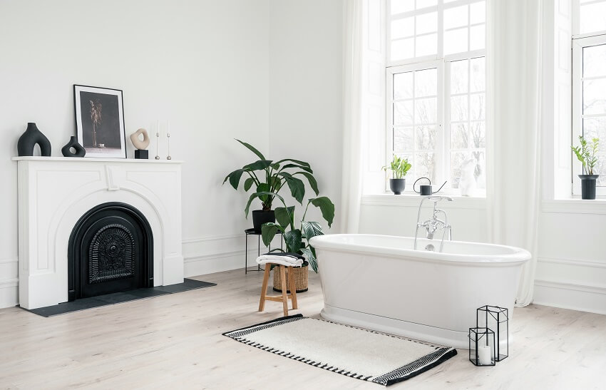 Белая ванная комната с отдельно стоящей фарфоровой ванной, комнатными растениями и декоративным камином