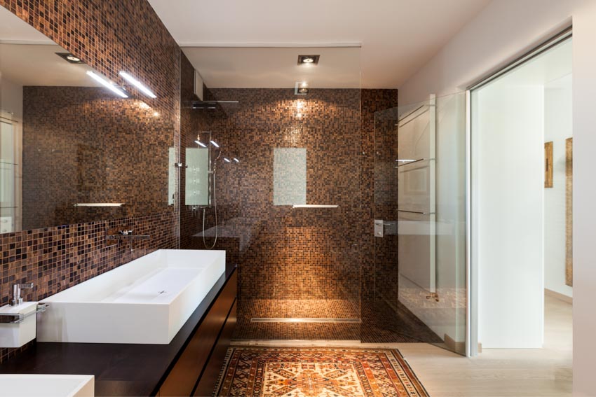 Красивая ванная комната с душем из стеклянной плитки, стеклянной дверью, напольным ковриком, раковиной, столешницей, зеркалом и потолочными светильниками.