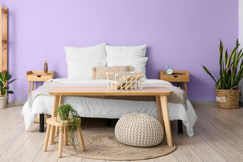 Спальня со стеной, окрашенной в лавандовый цвет, подушками, одеялом, деревянной скамейкой, оттоманкой, ковром, комнатным растением и стеной, окрашенной в лавандовый цвет.