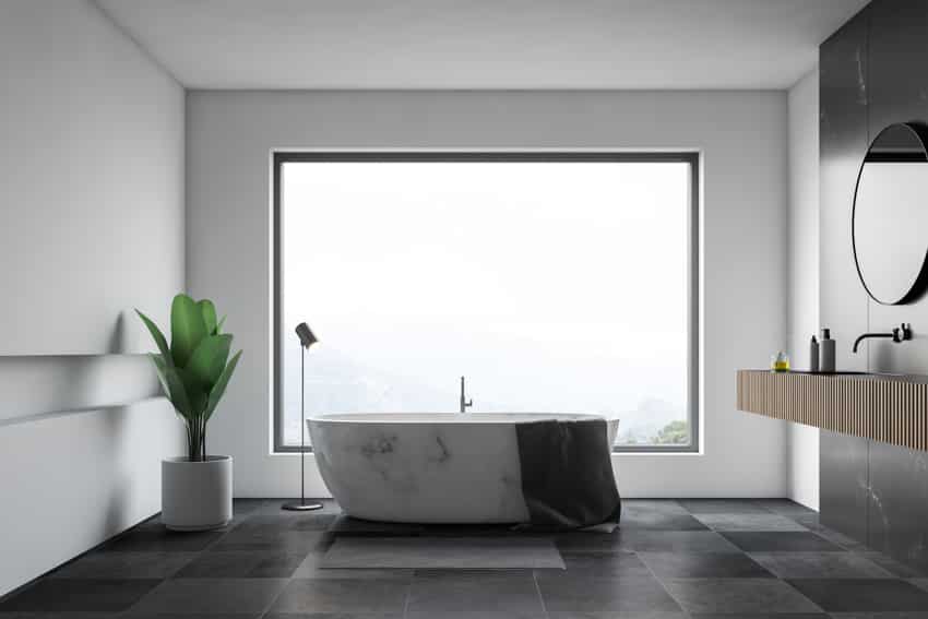 Ванная комната с панорамным окном, ванной из мраморного камня, кафельным полом, зеркальной раковиной и комнатным растением.