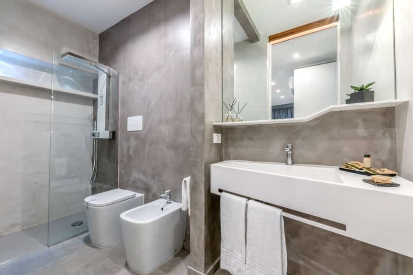 Ванная комната с бетонными стенами, кафельным полом, раковиной, зеркалом, двумя туалетами и душем.