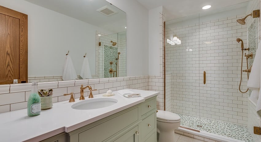 Светлая ванная комната с белой плиткой метро, ​​стеной, латунной фурнитурой, осветительными приборами, белыми столешницами, светло-зелеными ящиками и шкафами, а также мозаичным полом в душевой.