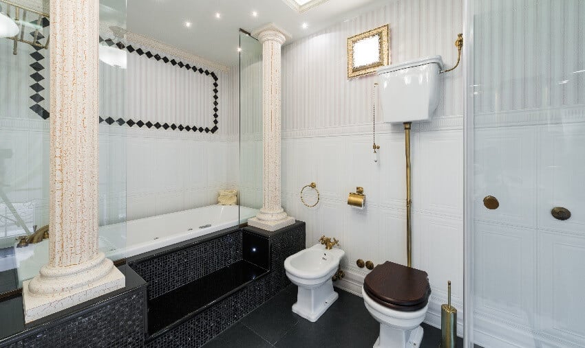 Современная ванная комната с колоннами, черная напольная плитка, ванна с черной мозаичной плиткой на ступенях, светильники и латунные держатели рулонов туалетной бумаги, кран для смыва и вешалка для полотенец.
