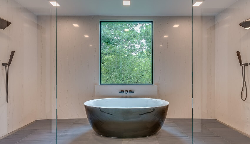 Роскошная современная ванная комната с отдельно стоящей ванной, душем на противоположных сторонах, большим окном и серым кафельным полом.