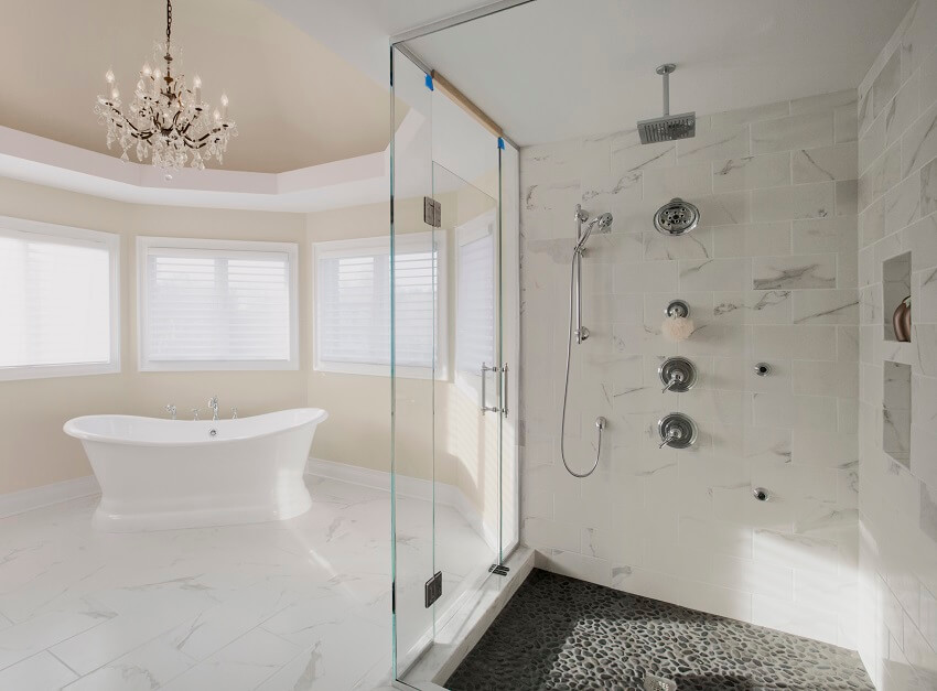 Большая главная ванная комната в жилом доме с двумя душевыми лейками, заключенными в стекло, с каменным полом, мраморной плиткой, стеной и отдельно стоящей ванной с люстрой наверху.