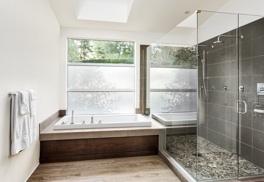 Большая ванная комната с ванной на деревянном полу и душем, заключенным в стекло, с двойным душем, мозаичным полом и темно-серой плиткой на стене.