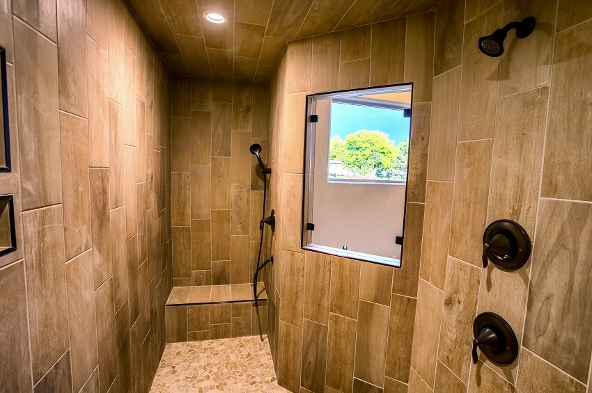 Жилая ванная комната со скамейкой, мозаичной плиткой, окном, двумя душевыми кабинами и деревянными стенами, выложенными плиткой.